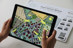Dieses Bild zeigt die "Smart Service Welten in AR"-App in Aktion auf einem Tablet. Im Hintergrund der für die App notwendige Augmented-Reality-Tracking-Flyer.
